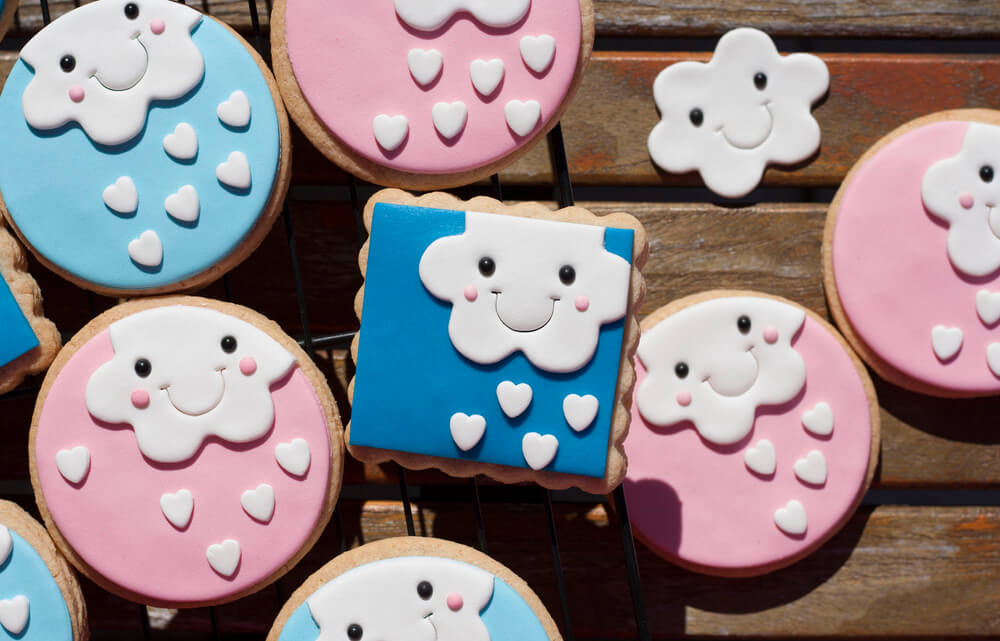 A imagem mostra biscoitos em formatos de quadrado e de círculo nas cores azul e rosa. Neles há o desenho de uma nuvem branca sorrindo e corações pequenos.