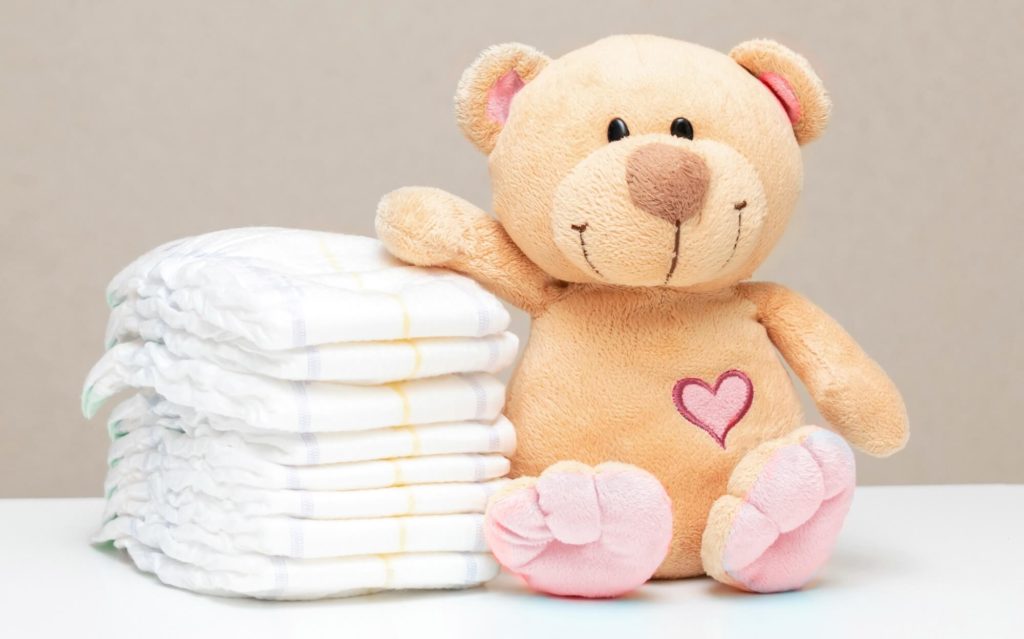  A imagem mostra várias fraldas em uma pilha ao lado de um urso de pelúcia. As fraldas são brancas e o urso é bege, com um coração rosa desenhado nele.