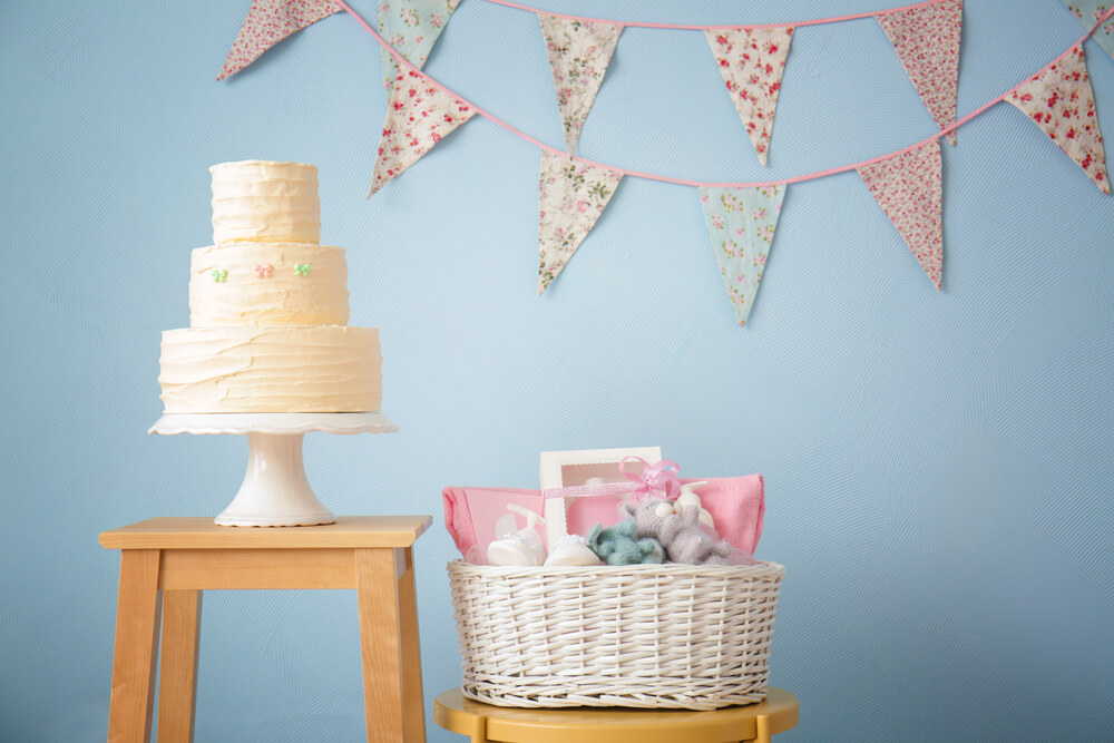 A imagem mostra uma decoração de festa de chá de bebê. Na imagem há um bolo de 3 camadas branco, um cesto de vime com vários presentes e bandeirolas ao fundo em uma parede azul.