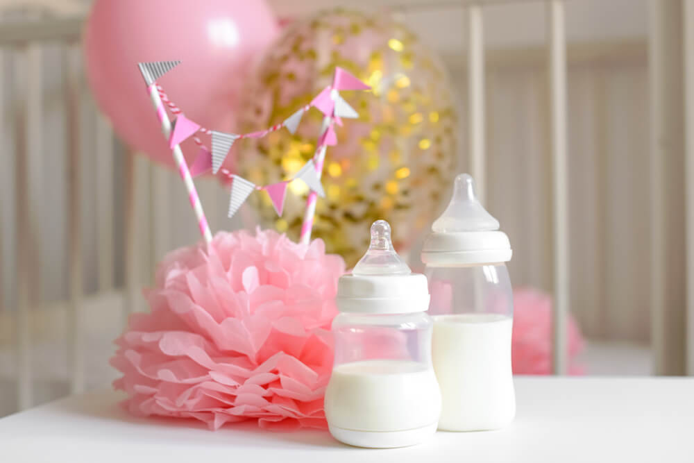 A imagem mostra uma decoração de chá de bebê com duas mamadeiras pequenas. Há também elementos de festa como balões e um enfeite rosa de papel.