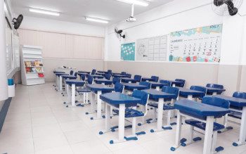 Foto que ilustra matéria sobre escolas particulares em Guarulhos mostra uma sala de aula da escola Mater Amabilis, em Guarulhos. A sala tem paredes brancas e chão claro. E as carteiras, conjuntos de cadeira e mesa, aparecem enfileiradas, lado a lado e uma em frente à outra. Elas têm estrutura branca de ferro e revestimento azul.