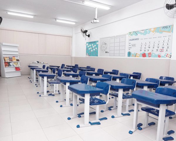 Foto que ilustra matéria sobre escolas particulares em Guarulhos mostra uma sala de aula da escola Mater Amabilis, em Guarulhos. A sala tem paredes brancas e chão claro. E as carteiras, conjuntos de cadeira e mesa, aparecem enfileiradas, lado a lado e uma em frente à outra. Elas têm estrutura branca de ferro e revestimento azul.