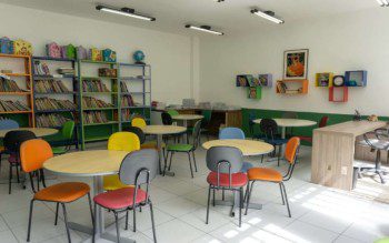 Foto que ilustra matéria sobre escolas em Osasco mostra uma sala de estudos do Colégio Cunha Carvalho, com diversas mesas redondas de madeira cercadas por cadeiras com estofados de diferentes cores. Ao fundo, há estantes com diversos livros.