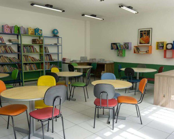 Foto que ilustra matéria sobre escolas em Osasco mostra uma sala de estudos do Colégio Cunha Carvalho, com diversas mesas redondas de madeira cercadas por cadeiras com estofados de diferentes cores. Ao fundo, há estantes com diversos livros.