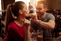 Foto que ilustra quiz de casal para o Dia dos Namorados mostra um homem dando um petisco na boca de uma mulher enquanto ela segura um copo de cerveja, representando um casal que gosta de comida de boteco