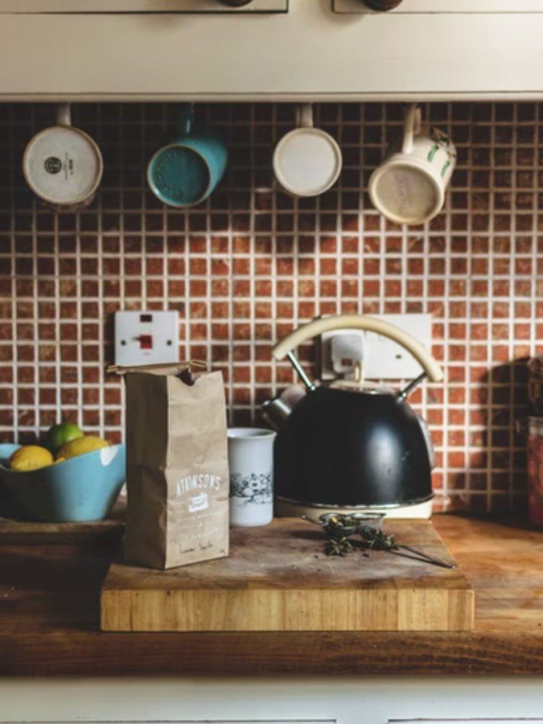 bancada de cozinha com vários utensílios, e armário na parede com canecas penduradas nele; a parede é azulejada