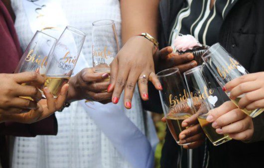 A imagem mostra as mãos de um casal de pessoas noivas e também de amigos ao redor deles. Todas as mãos seguram um copo com bebida, em sinal de comemoração.