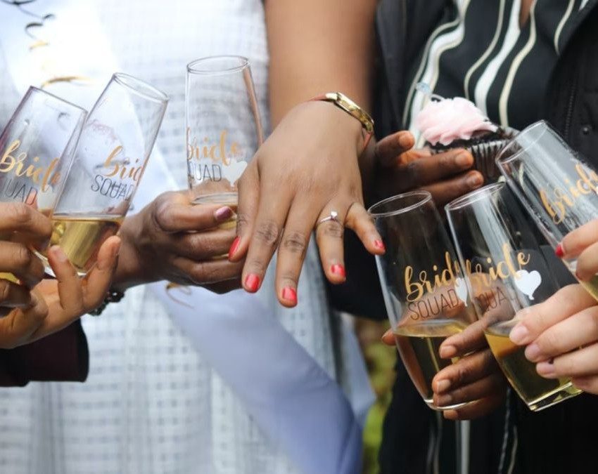 A imagem mostra as mãos de um casal de pessoas noivas e também de amigos ao redor deles. Todas as mãos seguram um copo com bebida, em sinal de comemoração.