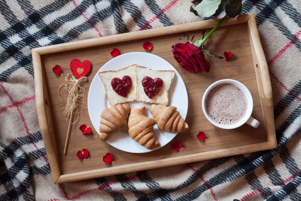 Tábua de madeira com café-da-manhã decorado com tema de dia dos namorados. Existem corações e pétalas de flores ao lado.