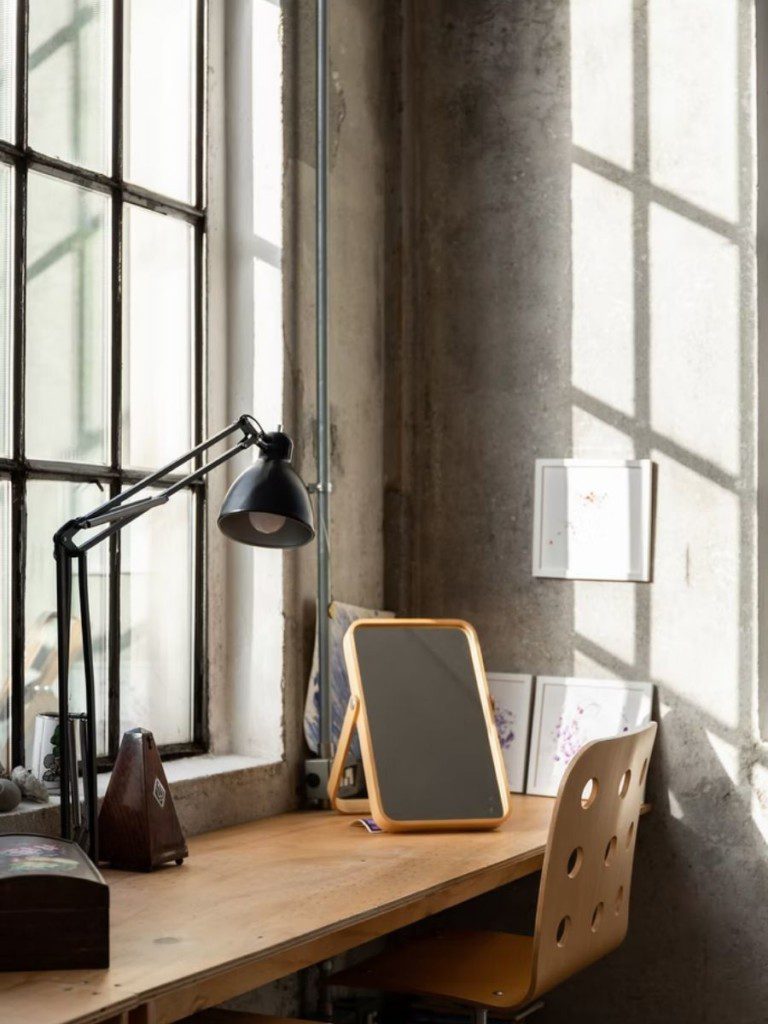 A imagem mostra uma mesa de trabalho com espelho e lâmpada em cima, encostada na parede com uma janela. A parede é feita de cimento queimado, dando um ar bem ao estilo industrial.