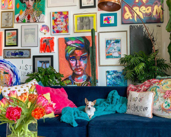 A foto mostra um exemplo de uma sala com decoração maximalista. Na parede há vários quadros de estilos e cores diferentes. O sofá e o puff são de cor azul escura. No sofá há várias almofadas e um cachorrinho enrolado em uma manta azul clara. No puff uma planta com flores e uma revista.