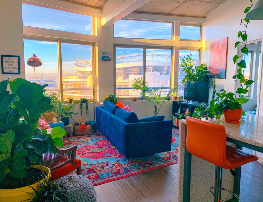 Foto de um ambiente aberto em um apartamento. Os principais elementos são plantas de diversas, um sofá azul escuro, uma cadeira laranja, um tapete bem colorido em tons de rosa e azul claro. Há também uma janela grande que ilumina o ambiente.