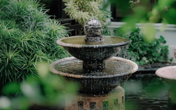 Imagem de uma fonte com água escoando e um jardim com bastante plantas verdes.