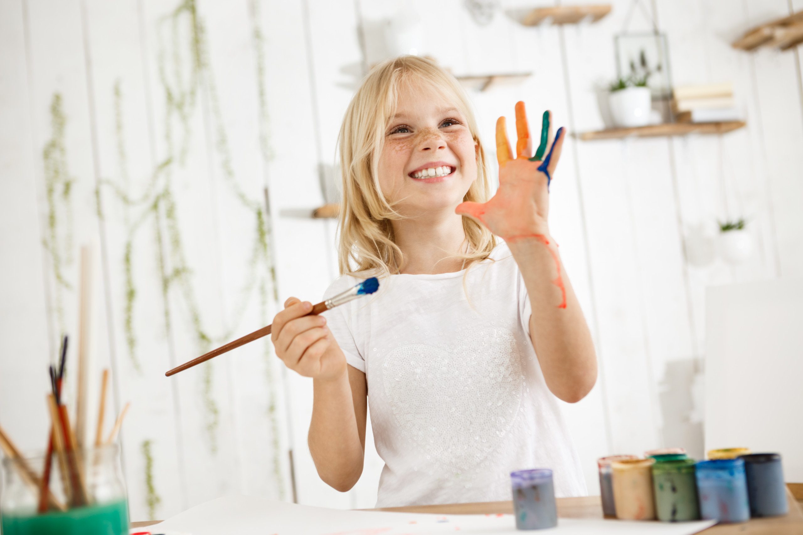 Imagem e uma menina brincando com tintas com um pincel na mão e a outra mão suja de tinta.