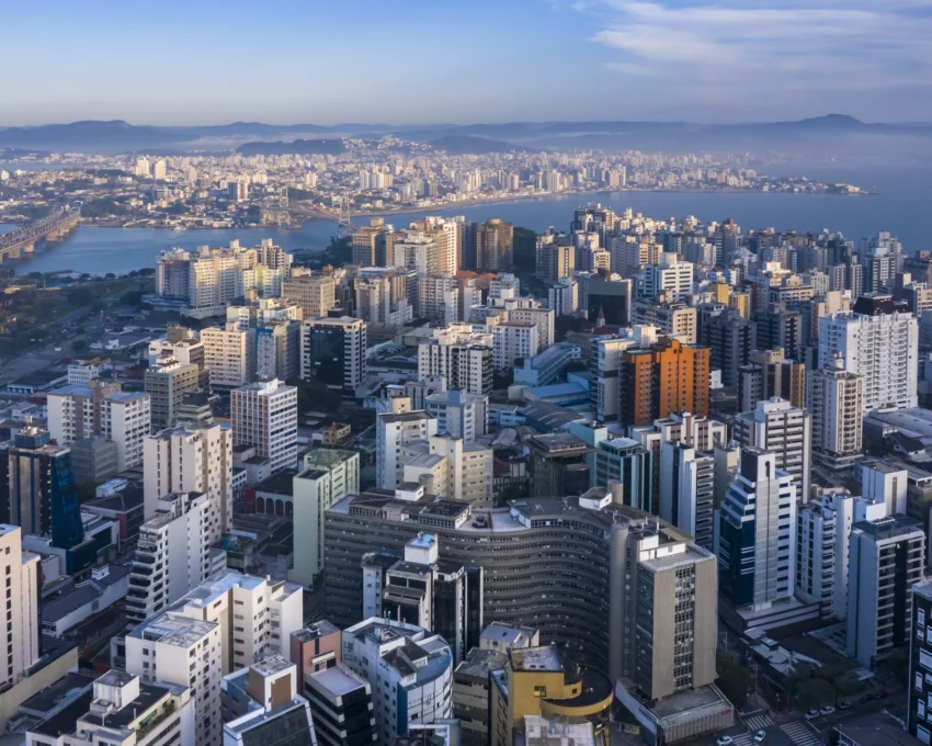 Imagem de edifícios do centro de Florianópolis vistos do alto com ponte suspensa a cabo ao fundo para ilustrar matéria sobre as melhores cidades de Santa Catarina
