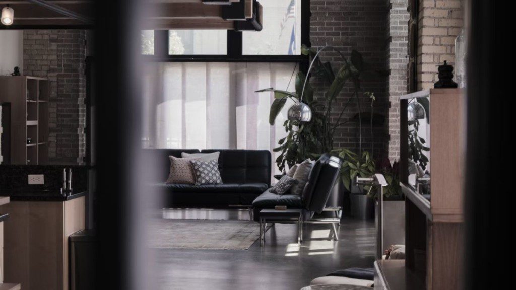 A imagem mostra um ambiente com sofá, plantas, armários e móveis em decoração estilo industrial, com janelas largas ao fundo