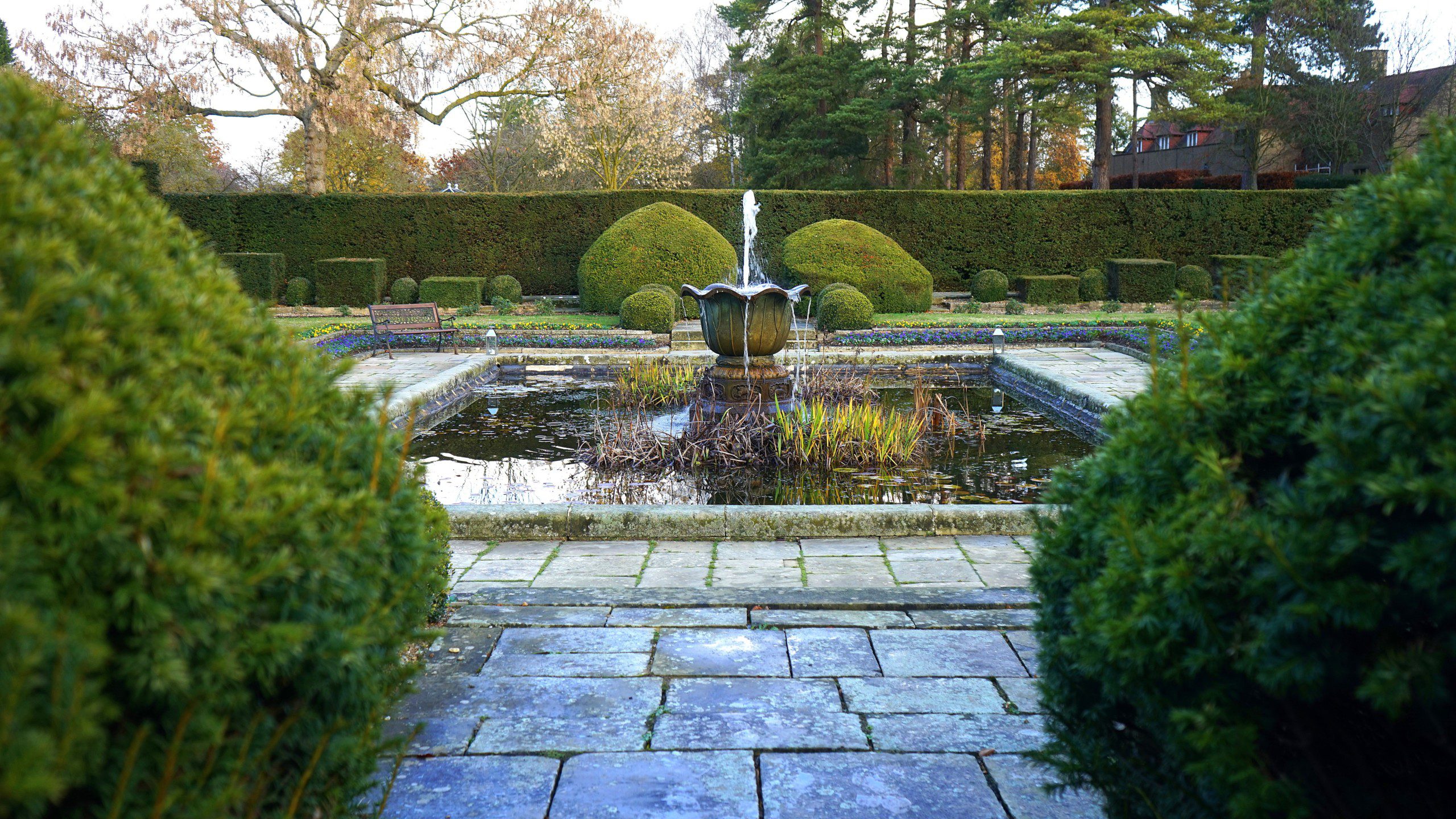  Imagem de uma fonte de água no centro de um jardim com bastante áreas verdes. 