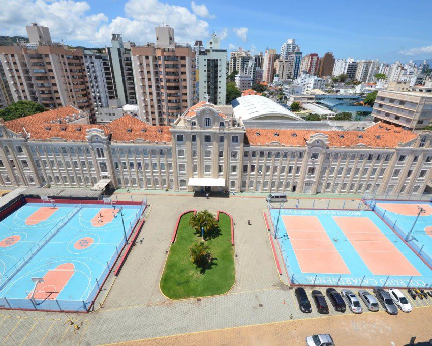 Foto que ilustra matéria sobre escolas particulares em Florianópolis mostra a fachada do Colégio Catarinense. À frente do prédio, há quadras poliesportivas. E atrás dele há prédios residenciais da cidade. Mais ao fundo, o céu azul de um dia ensolarado.