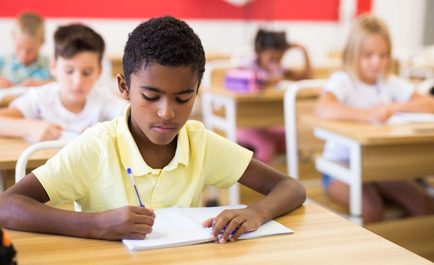 Foto que ilustra matéria sobre escolas em Americana mostra um menino negro, de camisa polo amarela, sentado a uma carteira de escola e fazendo anotações com uma caneta em um caderno.