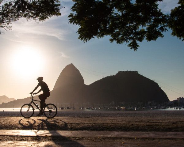 Foto que ilustra matéria sobre morar no Rio de Janeiro mostra a silhueta do morro do Pão de Açúcar. Ao lado esquerdo dele, aparece o sol baixo. E em primeiro plano, aparece a silhueta de um ciclista.