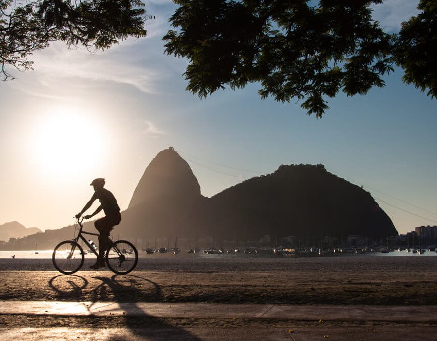 Foto que ilustra matéria sobre morar no Rio de Janeiro mostra a silhueta do morro do Pão de Açúcar. Ao lado esquerdo dele, aparece o sol baixo. E em primeiro plano, aparece a silhueta de um ciclista.