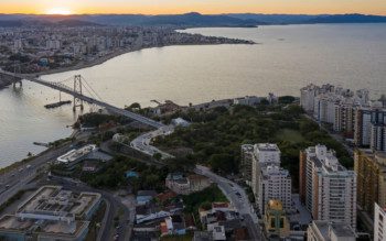 Foto que ilustra matéria sobre parques em Florianópolis mostra uma vista aérea do Parque da Luz. A imagem mostra área arborizada no centro da tela. Do meio para o fundo, um espelho d'água da baía. A ponte Hercílio Luz começa próximo à área arborizada. Na parte inferior da imagem, aparecem alguns prédios.