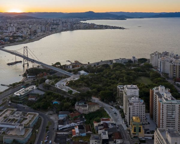 Foto que ilustra matéria sobre parques em Florianópolis mostra uma vista aérea do Parque da Luz. A imagem mostra área arborizada no centro da tela. Do meio para o fundo, um espelho d'água da baía. A ponte Hercílio Luz começa próximo à área arborizada. Na parte inferior da imagem, aparecem alguns prédios.