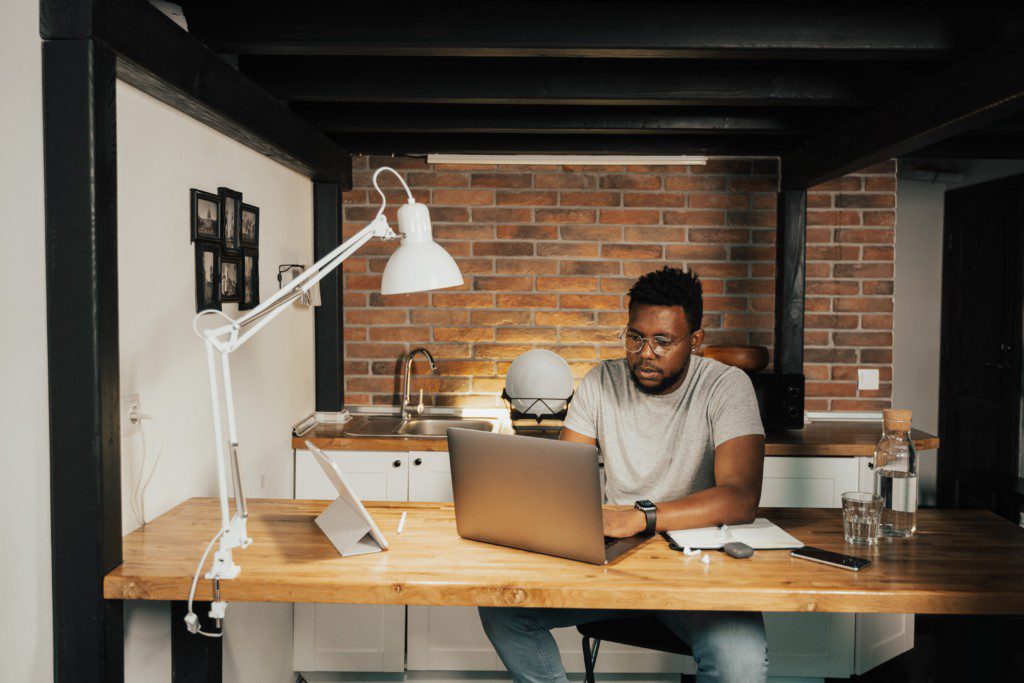 A foto mostra um escritório no estilo industrial com uma mesa ampla e parede de tijolos. Na mesa tem uma luminária em metal, tablet, notebook e um caderno de anotações. Um homem negro está sentado diante da mesa trabalhando no computador. 