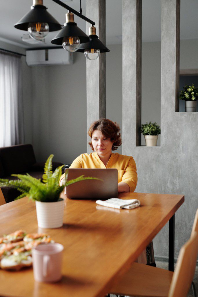 A foto mostra um escritório no estilo industrial com uma parede vazada em cimento, trilho de luzes e mesa de madeira. Há uma mulher sentada diante da mesa trabalhando em um notebook.