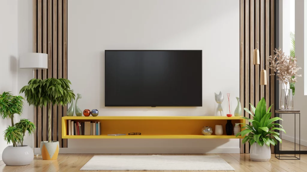 Imagem da decoração de uma sala de estar mostra rack suspenso na cor amarela com alguns livros e objetos decorativos em cima, plantas, um tapete branco e uma televisão pendurada na parede 