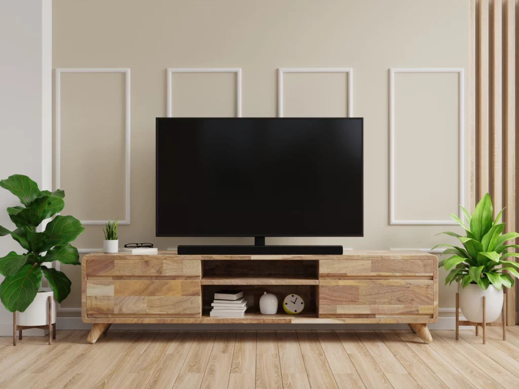 Imagem da decoração de uma sala de estar mostra rack de madeira, plantas, televisão, parede de cor creme e piso de madeira para ilustrar matéria sobre racks diferentes para sala