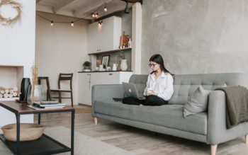 Foto de uma sala de estar moderna em tons de cinza e elementos em madeira. Na foto há uma mulher sentada no sofá cinza, há também um tapete e uma mesa de centro.