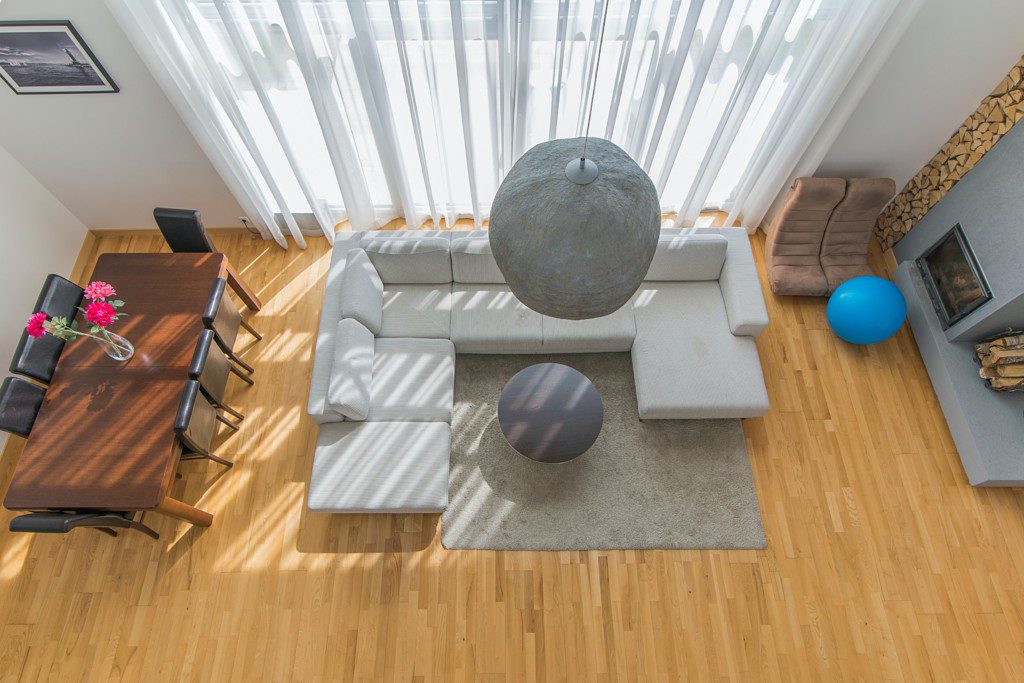 A foto mostra uma sala de estar moderna vista de cima. Nela há um sofá, um puff, um tapete e outros elementos secundários ao redor que compõem a casa.