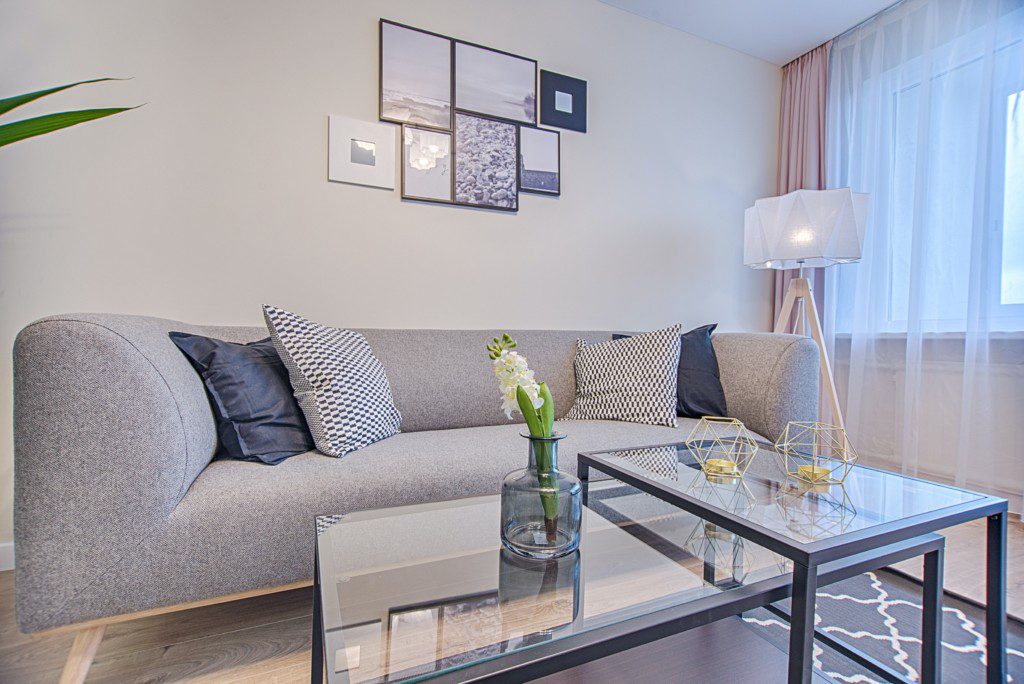 Foto de uma sala de estar moderna com sofá, quadros na parede e uma mesa de centro de metal e vidro. Uma luminária de chão agrega elegância ao ambiente em tons de branco, preto e cinza. 
