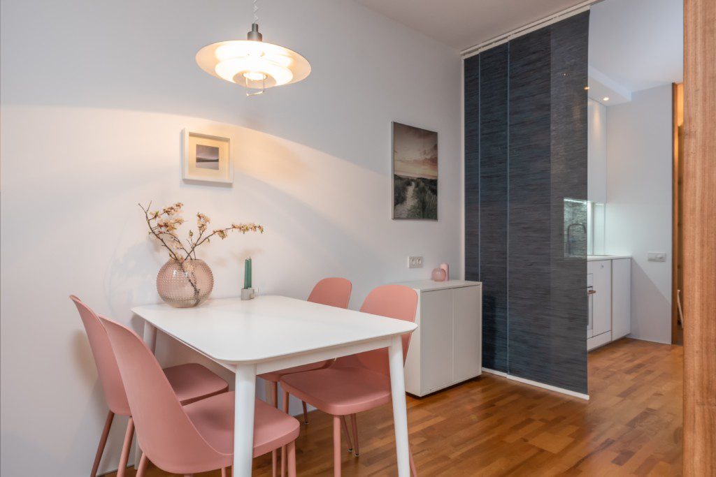 Foto de uma sala de jantar composta por: mesa com 4 cadeiras; aparador pequeno de duas portas; arranjo delicado na mesa e quadros decorativos bem discretos.