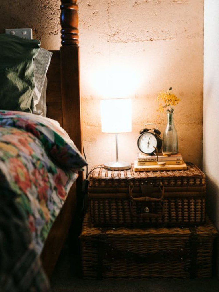 cama rústica ao lado de mesa de cabeceira, relógio pequeno antigo, vaso de flor e abajur antigo