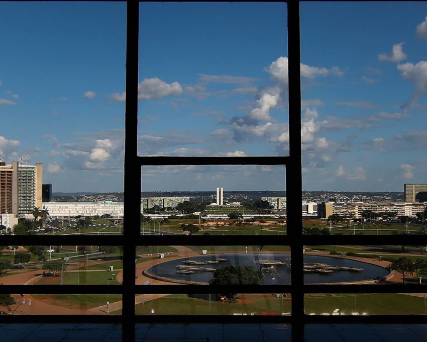Foto que ilustra matéria sobre bairros de Brasília mostra a Esplanada dos Ministérios vista de dentro de um prédio localizado de frente para a região. Em primeiro plano, aparecem, em sombras, a estrutura das janelas do prédio de onde a foto foi tirada. Bem ao fundo, aparece o Congresso Nacional.