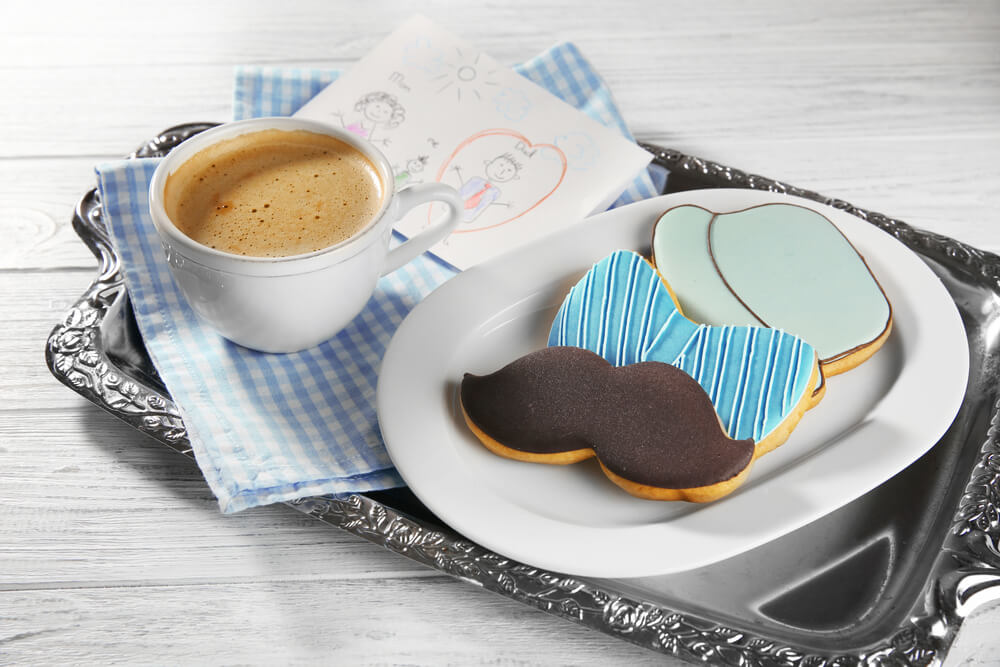 Foto que ilustra matéria sobre decoração para dia dos pais mostra uma bandeja de prata com uma xícara cheia de café, um desenho feito por crianças e biscoitos em forma de bigode, gravata borboleta e chapeu.