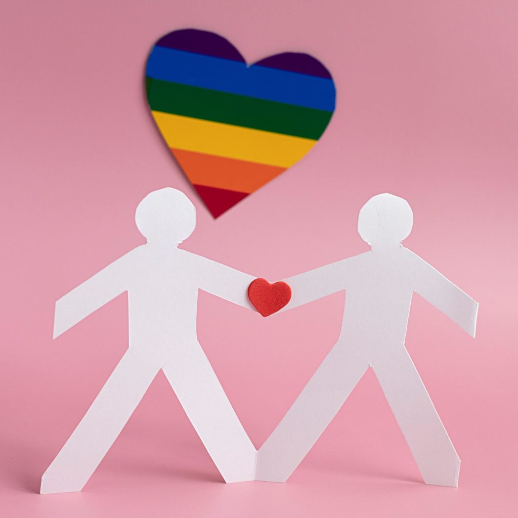 Foto que ilustra matéria sobre decoração para dia dos pais mostra dois bonecos de papel ligados pelas mãos por um coração vermelho. Ao fundo, um coração com as cores da bandeira LGBTQIAPN+