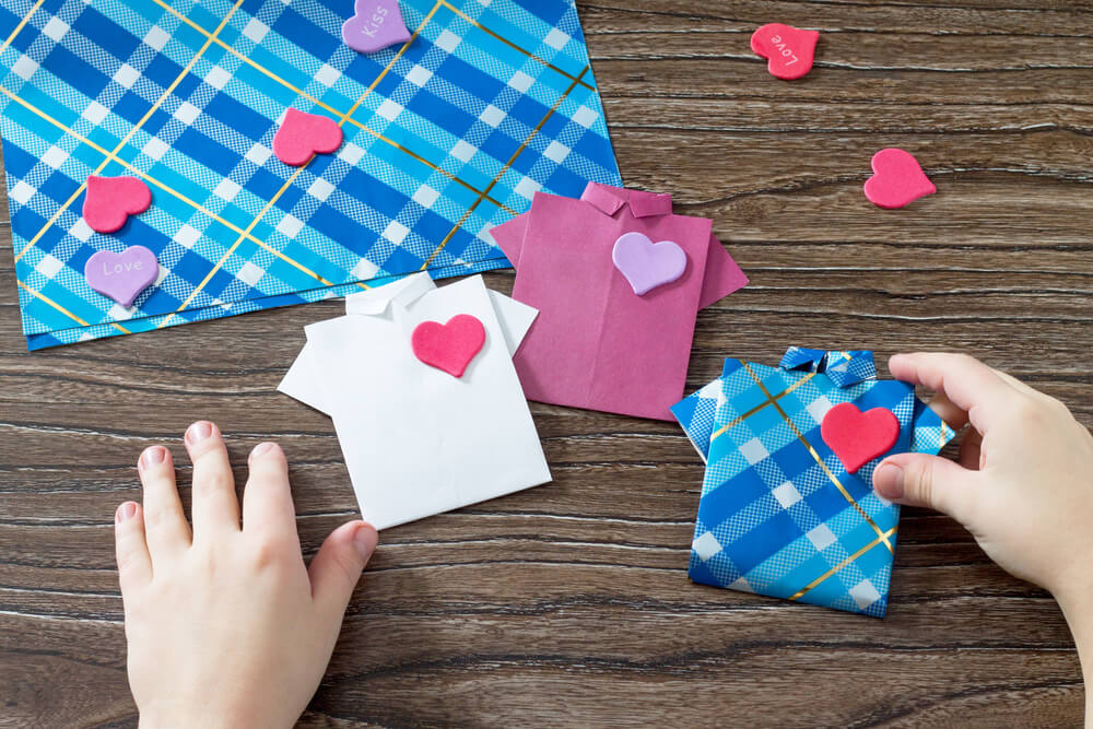 Foto que ilustra matéria sobre decoração para dia dos pais mostram as mãos de uma criança colando corações de EVA em pequenas réplicas de camisas sociais feitas de papel.