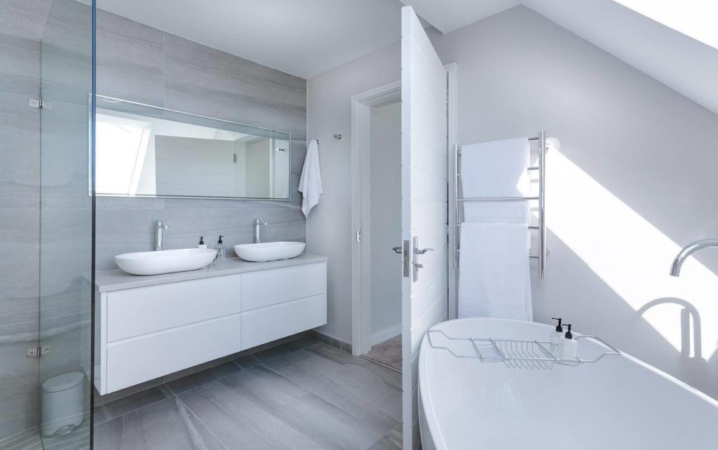 A imagem mostra um exemplo de banheiro minimalista. Nele há duas pias com armário embaixo delas, espelho retangular, banheira oval e uma área para banho. Tem também toalhas e itens de higiene pessoal.