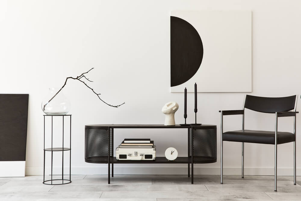 A imagem mostra uma sala de estar com uma estante pequena, um rack de ferro preto, uma cadeira e um quadro. 