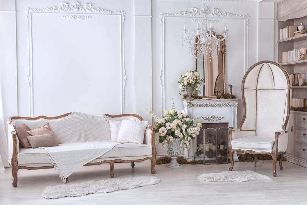  A imagem mostra um sofá, uma poltrona, espelho, estante de livros, dois tapetes e muitas flores em vasos. Tudo em um estilo clássico e bem requintado.
