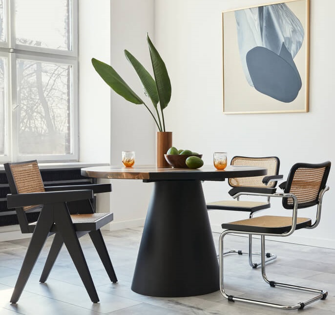 A imagem mostra uma sala de jantar com uma mesa redonda que possui três cadeiras ao redor. Há também um quadro e uma planta decorando o ambiente.