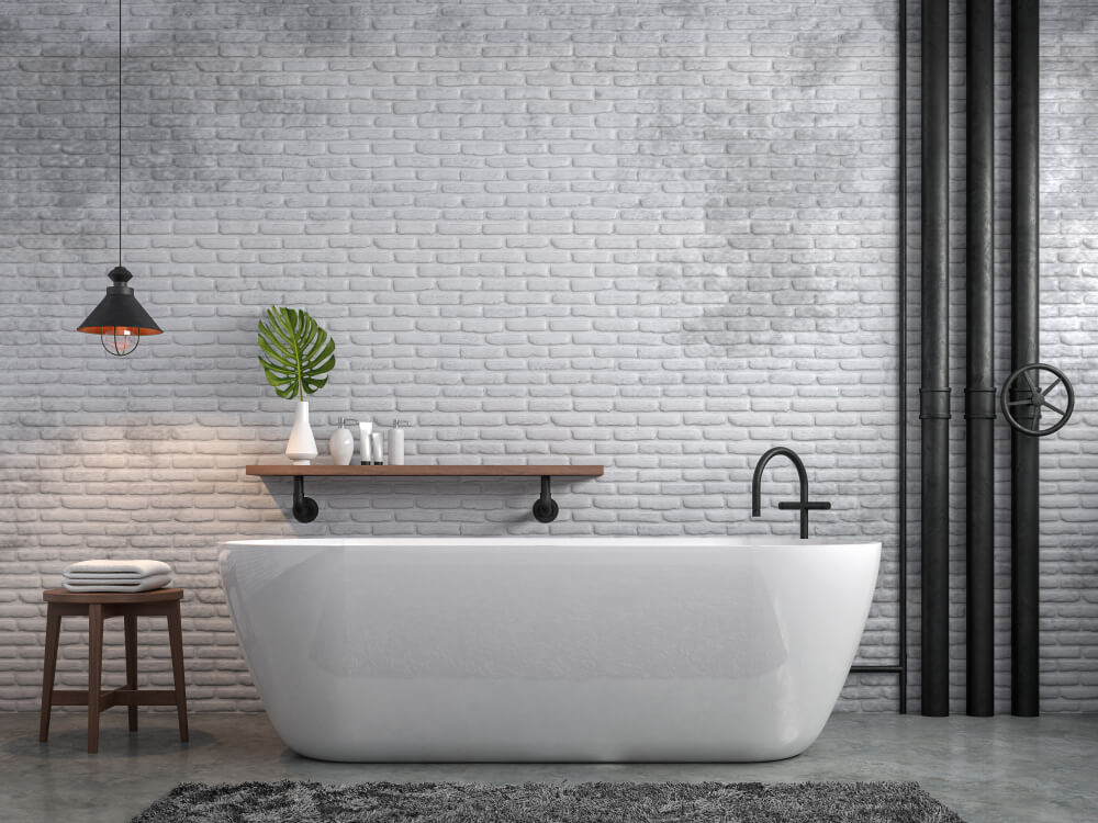 A imagem mostra um banheiro com uma banheira oval, um lustre de teto, um banquinho, um tapete e uma tubulação de ferro aparente na parede. Há também itens de higiene pessoal. 