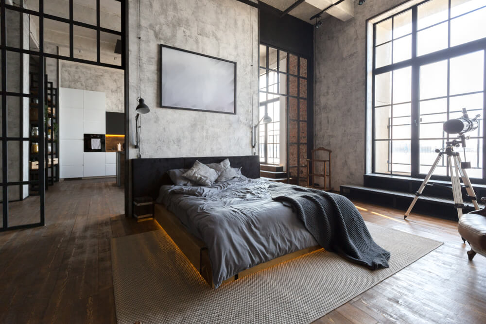 A imagem mostra um quarto com cama de casal, tapete, parede de cimento aparente. Há também uma luneta perto da janela.