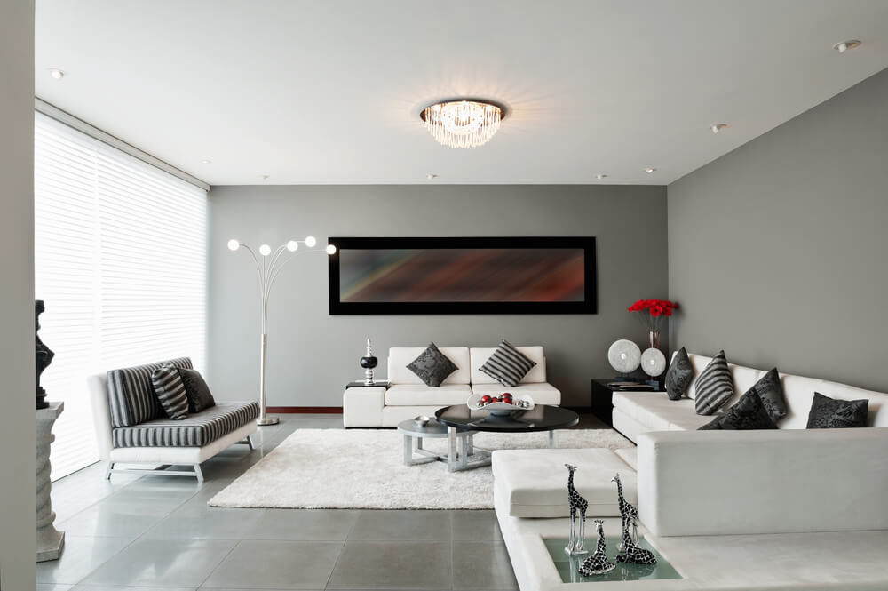 A imagem mostra uma sala de estar com três sofás, um amplo tapete, mesa de centro, além de decorações como um quadro, almofadas, luminária de chão.