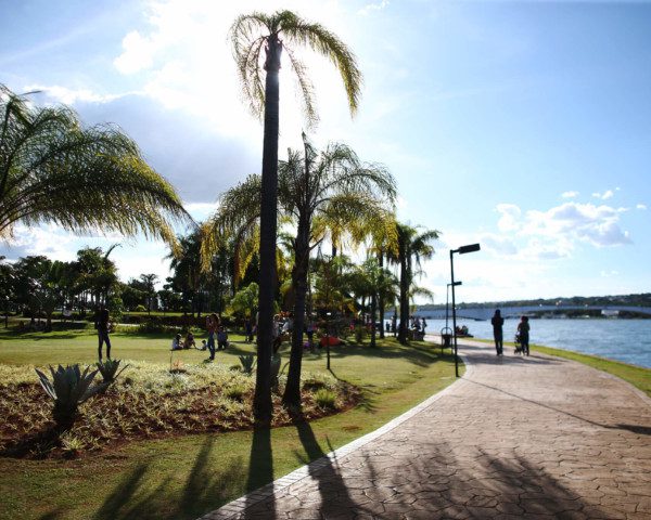 Foto que ilustra matéria sobre o que fazer em Brasília mostra a orla do Lago Paranoá, com o espelho d’água no canto direito, uma pista de caminhada no centro e um gramado com coqueiros à esquerda. Ao fundo, um céu azul ensolarado.