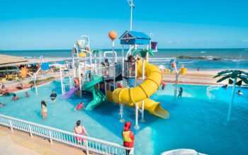 Foto que ilustra matéria sobre parque aquático em Salvador mostra o brinquedo aquático do Salvador Beach Club, uma estrutura com escorregadores coloridos em meio a uma piscina, com uma praia ao fundo.