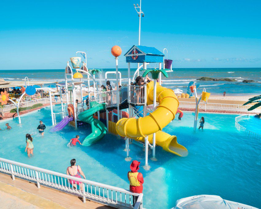 Foto que ilustra matéria sobre parque aquático em Salvador mostra o brinquedo aquático do Salvador Beach Club, uma estrutura com escorregadores coloridos em meio a uma piscina, com uma praia ao fundo.